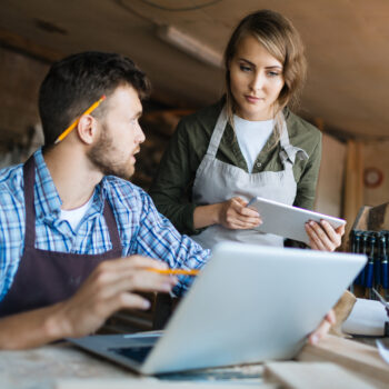 Pretty young carpenter and her bearded colleague brainstorming in workshop: he showing her something on computer screen while she looking at it with interest